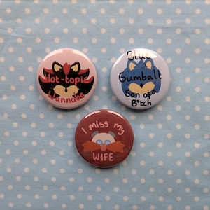 May include: Three round button badges featuring cartoon characters and text. The first badge is pink with a black and red cartoon character with the text "Hot-topic Wannabe". The second badge is blue with a blue cartoon character with the text "Blue Gumball Son of a B*tch". The third badge is brown with a white cartoon character with the text "I miss my WIFE".
