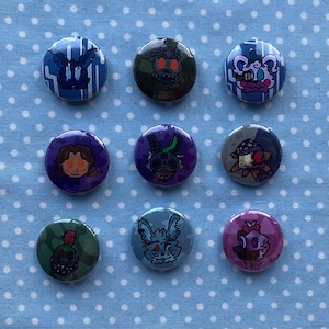 May include: Nine circular pin badges with cartoon character illustrations in various colours. The badges feature characters in shades of blue, green, purple, and grey. The badges are arranged on a light blue fabric with white polka dots.