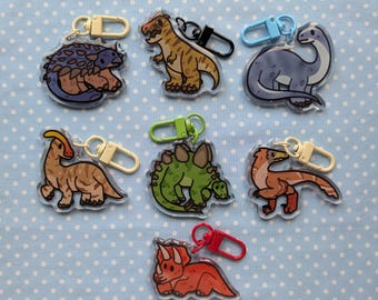 Könnte beinhalten: Acht bunte Dinosaurier-Schlüsselanhänger mit verschiedenen Designs. Jeder Schlüsselanhänger zeigt eine andere Dinosaurierart, darunter ein Stegosaurus, ein Triceratops, ein Brachiosaurus und ein Velociraptor. Die Schlüsselanhänger sind aus klarem Acryl gefertigt und haben einen Metallring zum Anbringen an Schlüsseln oder Taschen.
