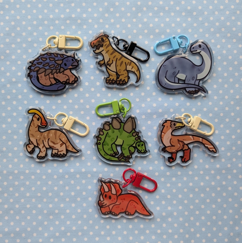May include: Eight colorful dinosaur keychains with different designs. Each keychain features a different dinosaur species, including a stegosaurus, a triceratops, a brachiosaurus, and a velociraptor. The keychains are made of clear acrylic and have a metal ring for attaching to keys or bags.