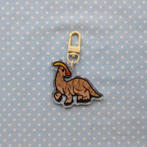 May include: A brown and white dinosaur keychain with a yellow crest. The keychain is made of clear acrylic and has a silver metal ring.