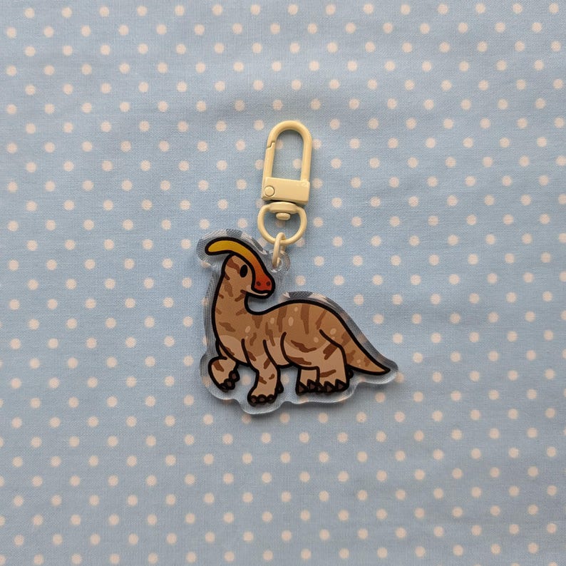 May include: A brown and white dinosaur keychain with a yellow crest. The keychain is made of clear acrylic and has a silver metal ring.