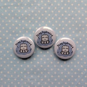 May include: Three circular, pale blue badges with the words "Stay hydrated" in black. Each badge displays a smiling cartoon character with a blue cloud and water droplet design. The badges are set against a light blue polka dot backdrop.