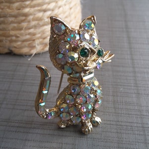 May include: A gold-tone brooch in the shape of a cat, adorned with iridescent rhinestones and green glass eyes. The cat has a raised tail and a small collar.