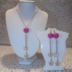 Op de afbeelding: Een sieradenset bestaande uit een goudkleurige ketting en bijpassende oorbellen. Elk sieraad is voorzien van een gladde, ronde, magentakleurige kraal, een kleine witte vlinderbedel en een witte bloembedel aan fijne goudkleurige kettinkjes.