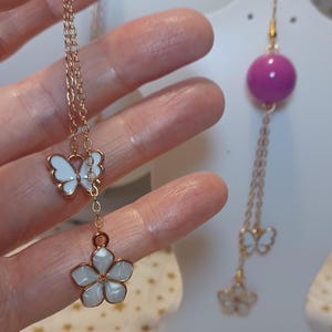Op de afbeelding: Een paar goudkleurige bungelende oorbellen met een paarse bolvormige kraal aan de bovenkant, gevolgd door een gouden ketting met een witte emaille vlinderbedel en een witte emaille bloembedel met vijf bloemblaadjes aan de onderkant.
