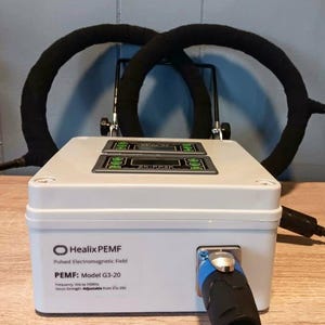 May include: A white HealiX PEMF device with two black, circular coils. The device has two digital displays and a blue and black connector. Text on the device reads "HealiX PEMF" and "PEMF: Model G3-20".