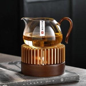 May include: A clear glass teapot with a wooden base and a metal heating element. The teapot has a handle and a spout. The base has a decorative design.