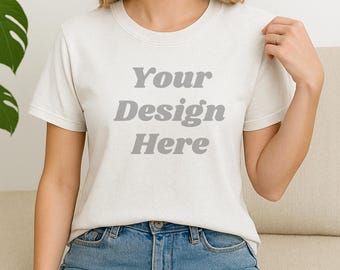 May include: A light-colored t-shirt with the text "Your Design Here" in gray block letters. The shirt is being worn with blue jeans. The model is sitting on a beige couch.