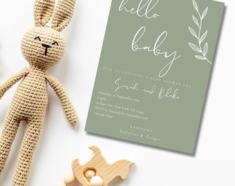 Könnte beinhalten: Eine Babyparty-Einladung mit dem Schriftzug "hello baby" in weißer Schrift auf einem salbeigrünen Hintergrund. Ein gehäkeltes Häschen und ein Holzspielzeug liegen neben der Einladung. Die Einladung enthält Details für die Party.