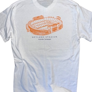Puede incluir: Camiseta blanca con un dibujo naranja de contorno del estadio Neyland, hogar del equipo de fútbol americano de los Volunteers de la Universidad de Tennessee. El texto "Neyland Stadium" y "Knoxville, Tennessee" está impreso debajo del estadio.