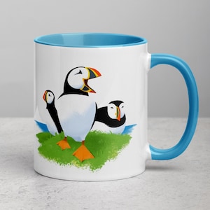 May include: White ceramic mug with a light blue rim and handle. The mug features a colorful illustration of three puffins on a green grassy island.