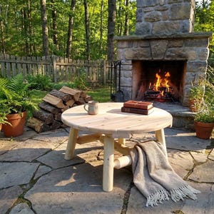 May include: A round, light-colored wooden coffee table with four legs sits on a stone patio. A mug and two books rest on the table. A fringed blanket is draped over the side. A stone fireplace with a burning fire is in the background.