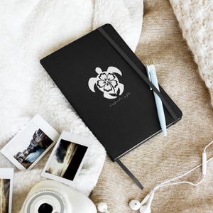 May include: Black notebook with a white turtle and flower design. The notebook has a black elastic band and a matching ribbon bookmark. A light blue pen rests on the notebook. The text "wayfinder gifts" is printed below the turtle design.