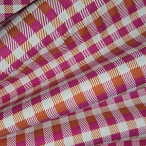 May include: Close-up of a fabric with a gingham pattern. The fabric features a repeating pattern of squares in white, fuchsia, and orange. The fabric is folded, creating shadows and depth, suggesting it is a textile for sewing or crafting.