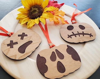 May include: Three wooden pumpkin ornaments with carved faces and orange ribbon hangers. The pumpkins are light brown with dark brown cut-out features. A yellow sunflower and orange leaves are in the background, suggesting a fall theme.