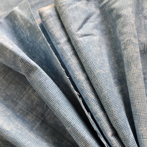 May include: Close-up of folded fabric in shades of blue and white, with a subtle grid pattern. The fabric has a soft, textured appearance, and the edges are finished with a light-colored trim. The folds create a fan-like arrangement.