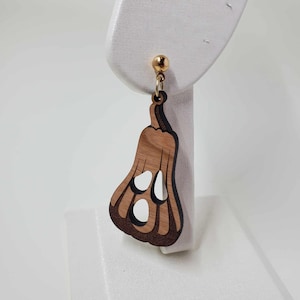 Könnte beinhalten: Ein einzelner ohrring in Kürbisform aus Holz mit einem goldfarbenen Stecker. Der Kürbis hat eine hellbraune Mitte und eine dunklere braune Umrandung mit drei ausgeschnittenen Löchern für Augen und Mund. Der Ohrring hängt an einer weißen Oberfläche.