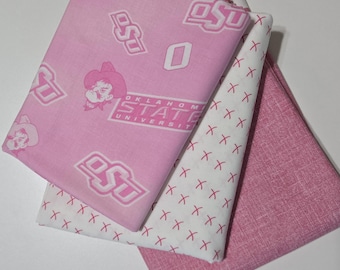 May include: Three folded pieces of fabric. One is light pink with white Oklahoma State University logos and mascot graphics. Another is white with a repeating pattern of small pink X marks. The third is a solid, textured dusty pink fabric.
