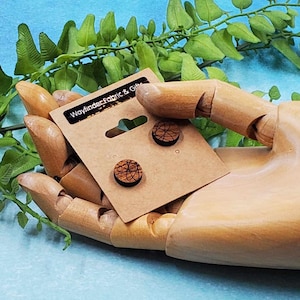 May include: A pair of wooden earrings with a black and brown geometric design. The earrings are on a brown card with the text "Wayfinder Fabric & Gifts".