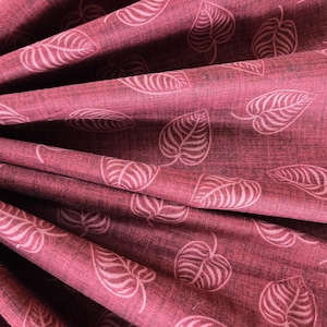May include: Close-up of a folded fabric with a burgundy background and a repeating pattern of white leaf designs. The fabric has a textured appearance, with the leaves varying in size and shape.