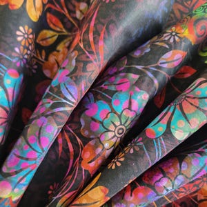 May include: Close-up of a fabric with a vibrant floral pattern. The design features colourful flowers in shades of orange, pink, blue, and green against a dark background. The fabric is folded, showcasing the intricate details of the print.