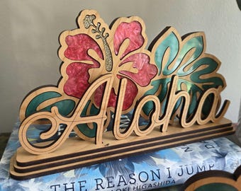 Puede incluir: Un letrero decorativo de madera con la palabra Aloha en letra cursiva, superpuesto sobre un recorte de flor de hibisco con pétalos de resina rosa brillante y estambre dorado, junto a hojas de resina verde sobre una base de madera.