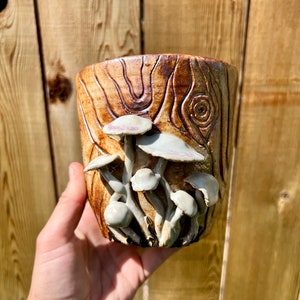 May include: A ceramic planter shaped like a tree trunk with a detailed wood grain texture. The planter is decorated with white and pink mushrooms.