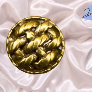 May include: A gold-colored metal button with a woven design. The button is round and has a raised, textured surface.
