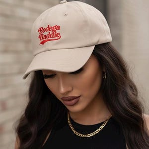 Op de afbeelding: Een beige baseballpet met de geborduurde tekst "Bodega Baddie" in rood schrift. De pet heeft een gebogen klep en een klassiek zes-panelen ontwerp. De persoon draagt lang, donker haar en een gouden ketting.