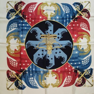 K&ouml;nnte beinhalten: Ein quadratisches Seidentuch mit einem dunkelblauen Mittelkreis und einem hellblauen Schiffsbug-Motiv. Die Ecken zeigen rote und blaue Schiffsbug-Motive mit goldenen Ankern. Der Text PROUES ist mittig auf einem wei&szlig;en Hintergrund gedruckt.