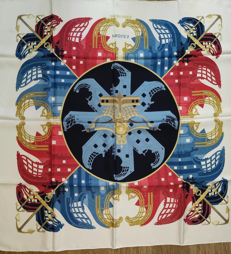 K&ouml;nnte beinhalten: Ein quadratisches Seidentuch mit einem dunkelblauen Mittelkreis und einem hellblauen Schiffsbug-Motiv. Die Ecken zeigen rote und blaue Schiffsbug-Motive mit goldenen Ankern. Der Text PROUES ist mittig auf einem wei&szlig;en Hintergrund gedruckt.