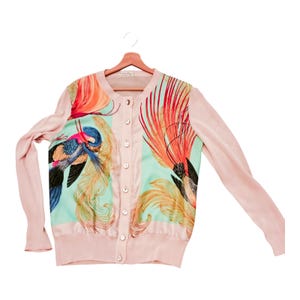 Peut inclure: Un cardigan rose clair à manches longues et à boutons. Les panneaux avant présentent un fond turquoise avec un motif coloré d'oiseaux et de plumes dans les tons orange, jaune, bleu et noir. Le cardigan est suspendu à un cintre en bois.