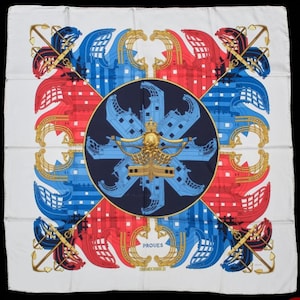 K&ouml;nnte beinhalten: Ein quadratisches Seidentuch mit wei&szlig;em Hintergrund und einem zentralen dunkelblauen Kreis, der ein goldenes Kronen- und Schiffsmotiv enth&auml;lt. Das Design zeigt stilisierte rote und blaue Schiffsr&uuml;mpfe und goldene Anker mit dem Text PROUES.