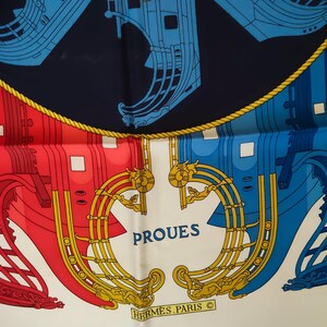 K&ouml;nnte beinhalten: Ein Seidenschal mit nautischem Design, das stilisierte Schiffsr&uuml;mpfe in Rot, Blau und Gold vor einem dunkelblauen und wei&szlig;en Hintergrund zeigt. Der Schriftzug PROUES ist in Blau und HERMES PARIS in einem goldenen Rechteck aufgedruckt.