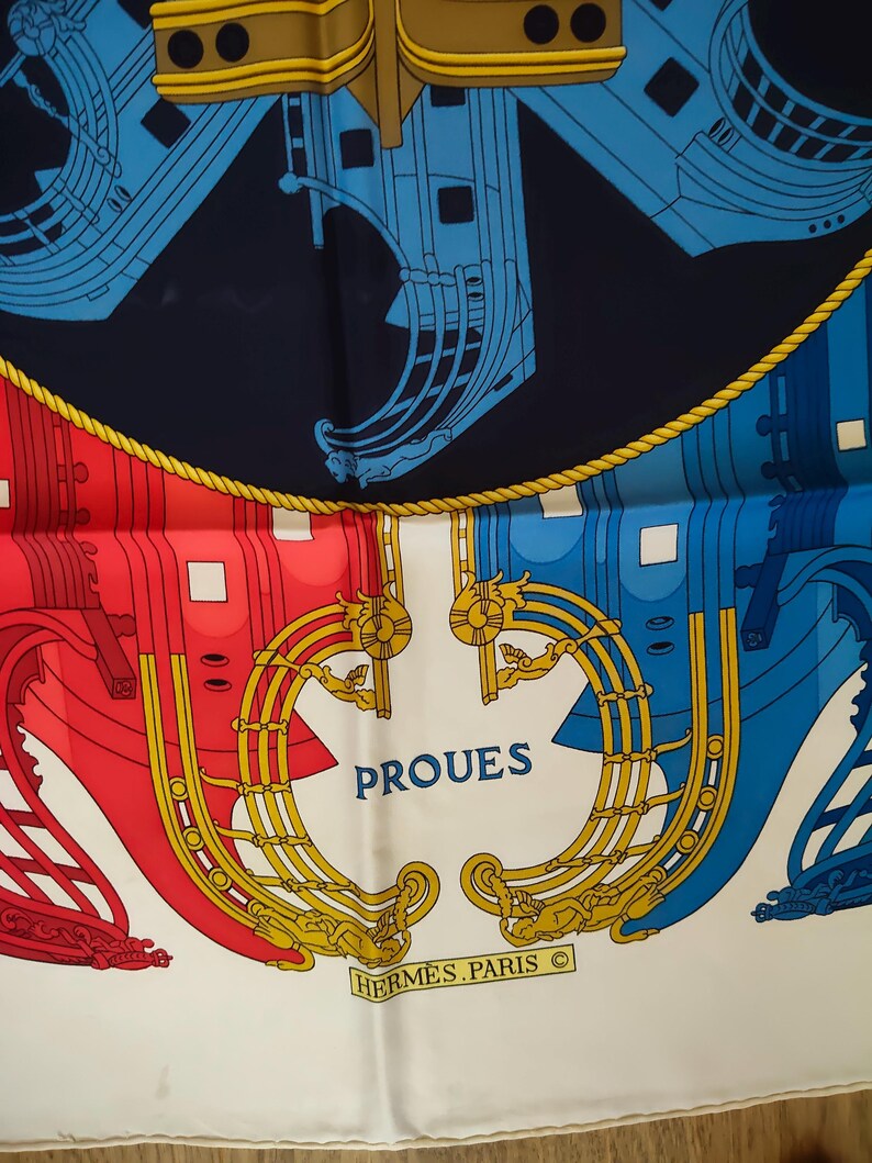 K&ouml;nnte beinhalten: Ein Seidenschal mit nautischem Design, das stilisierte Schiffsr&uuml;mpfe in Rot, Blau und Gold vor einem dunkelblauen und wei&szlig;en Hintergrund zeigt. Der Schriftzug PROUES ist in Blau und HERMES PARIS in einem goldenen Rechteck aufgedruckt.