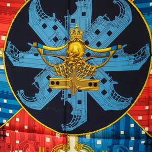K&ouml;nnte beinhalten: Ein quadratisches Seidentuch mit einem dunkelblauen Mittelkreis, der ein goldenes Kronen- und Schiffsbug-Emblem zeigt. Hellblaue Schiffsr&uuml;mpfe sind sternf&ouml;rmig angeordnet. Die Ecken zeigen geometrische Schiffsrumpfmuster in Rot und Blau.