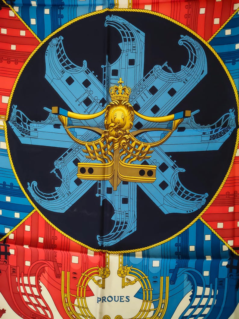 K&ouml;nnte beinhalten: Ein quadratisches Seidentuch mit einem dunkelblauen Mittelkreis, der ein goldenes Kronen- und Schiffsbug-Emblem zeigt. Hellblaue Schiffsr&uuml;mpfe sind sternf&ouml;rmig angeordnet. Die Ecken zeigen geometrische Schiffsrumpfmuster in Rot und Blau.