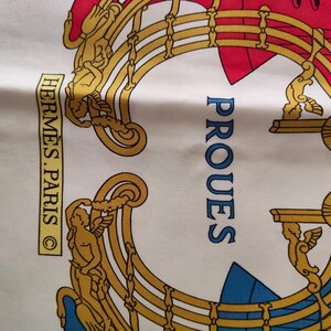K&ouml;nnte beinhalten: Ein wei&szlig;er Seidenschal mit einem goldfarbenen, verzierten Rahmendesign und gefl&uuml;gelten Figuren. Der Text HERM&Egrave;S.PARIS ist in einem gelben Rechteck gedruckt und das Wort PROUES ist in Blau gehalten. Das Design enth&auml;lt rote und blaue Akzente.