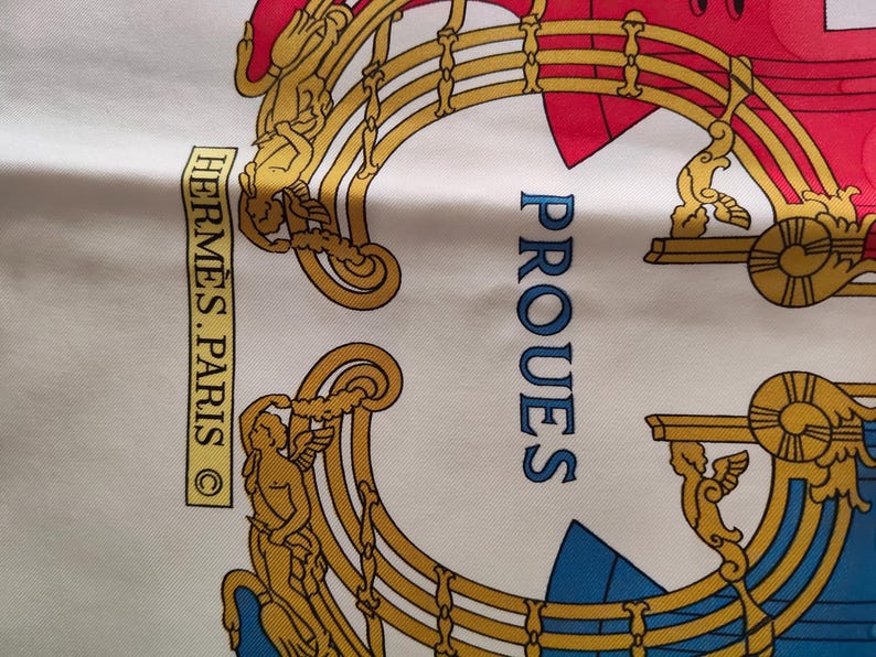 K&ouml;nnte beinhalten: Ein wei&szlig;er Seidenschal mit einem goldfarbenen, verzierten Rahmendesign und gefl&uuml;gelten Figuren. Der Text HERM&Egrave;S.PARIS ist in einem gelben Rechteck gedruckt und das Wort PROUES ist in Blau gehalten. Das Design enth&auml;lt rote und blaue Akzente.