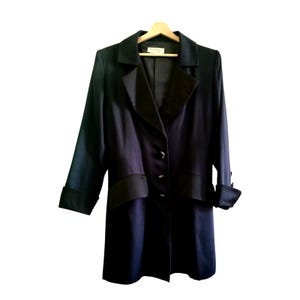 May include: A dark navy blue coat with black lapels and cuffs, hanging on a wooden hanger. The coat has a classic design with three buttons down the front and a satin-like finish.