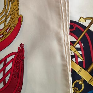 K&ouml;nnte beinhalten: Ein wei&szlig;er Seidenschal mit einem detaillierten nautischen Druck. Das Design zeigt links einen roten Schiffsrumpf mit goldenen Akzenten und rechts einen dunkelblauen Anker mit goldenen Seilen. Der Stoff hat eine glatte Textur mit einem sichtbaren Saum.