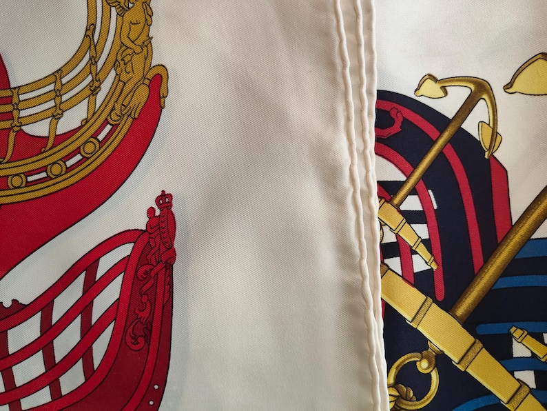 K&ouml;nnte beinhalten: Ein wei&szlig;er Seidenschal mit einem detaillierten nautischen Druck. Das Design zeigt links einen roten Schiffsrumpf mit goldenen Akzenten und rechts einen dunkelblauen Anker mit goldenen Seilen. Der Stoff hat eine glatte Textur mit einem sichtbaren Saum.