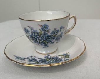 May include: A white teacup and saucer set with a floral pattern of blue flowers and green leaves. The rims are trimmed with gold. The teacup has a handle and sits on a matching saucer.