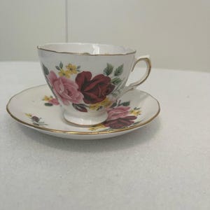 May include: A white teacup and saucer set featuring a floral rose pattern in pink and burgundy, with yellow accents and green leaves. The edges are trimmed with gold.