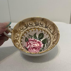 Może przedstawiać: Zabytkowy filiżanka do herbaty z różowym wzorem róży. Filiżanka ma złoty brzeg i uchwyt, ze złotymi akcentami filigranowymi wokół krawędzi. Wnętrze przedstawia dużą różową różę z zielonymi liśćmi.