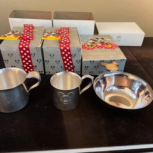 May include: Three silver baby cups and a silver bowl with Mickey Mouse designs. The cups and bowl are on a dark brown surface. The cups have handles and the bowl is round. The cups and bowl are all shiny and new. There are three boxes in the background with Mickey Mouse designs and red bows.