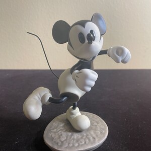 May include: Black and white porcelain figurine of Mickey Mouse in a running pose. Mickey is wearing his signature red shorts and white gloves. He is standing on a gray base with a textured surface.