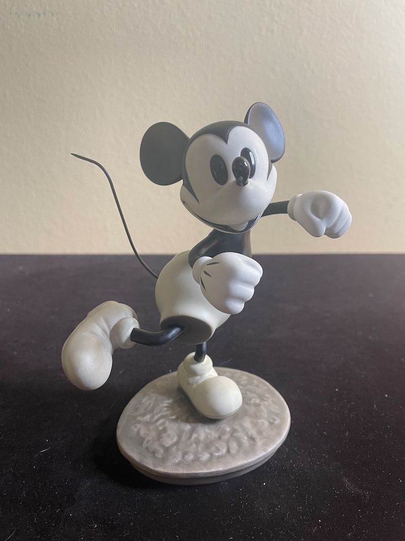 May include: Black and white porcelain figurine of Mickey Mouse in a running pose. Mickey is wearing his signature red shorts and white gloves. He is standing on a gray base with a textured surface.