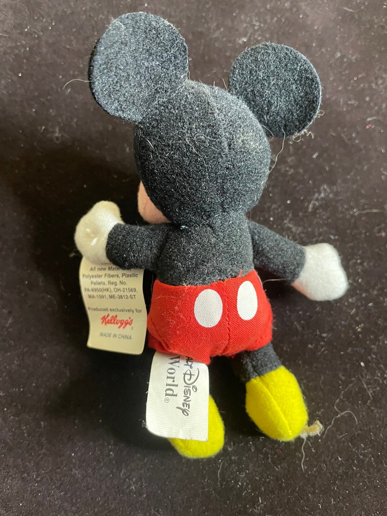 K&ouml;nnte beinhalten: Ein kleines schwarzes und rotes Pl&uuml;sch-Mickey-Mouse-Spielzeug mit gelben Schuhen. Das Spielzeug hat ein Etikett mit der Aufschrift "Disney World" und "Kellogg's".